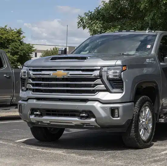 A silver Chevrolet pickup truck is parked in a lot on a sunny day, with trees and another vehicle visible in the background.
