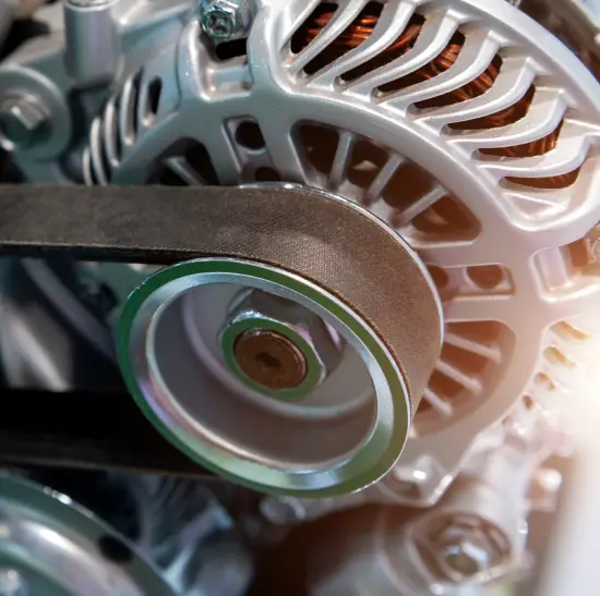Close-up of a car engine alternator with a black serpentine belt running over a shiny metal pulley, showing detailed ventilation slots and copper wiring inside.