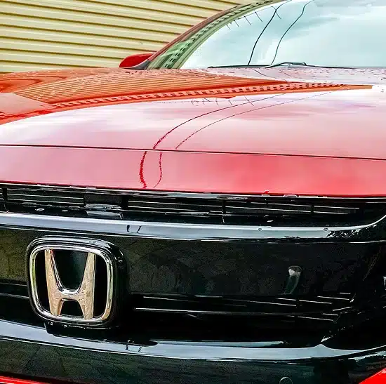 Close-up of the front of a red Honda car, showing the Honda emblem on the black grille with reflections visible on the hood and background.