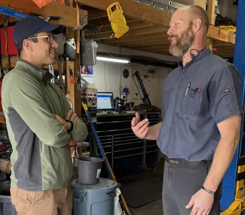 Auto repair in Tampa FL. SPES Automotive. Two men stand and talk inside a workshop or garage. One man wears glasses, a cap, and a green jacket, while the other wears a work shirt and has a beard. Tools and equipment are visible in the background.