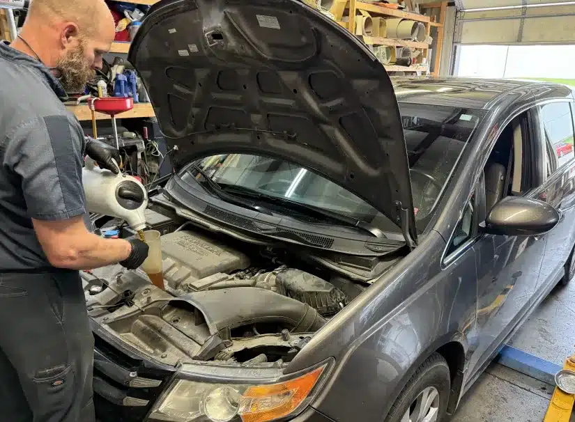 Preventative Maintenance at SPES automotive. A mechanic pours oil into the engine of a gray minivan with its hood open inside an auto repair shop filled with various tools and equipment.