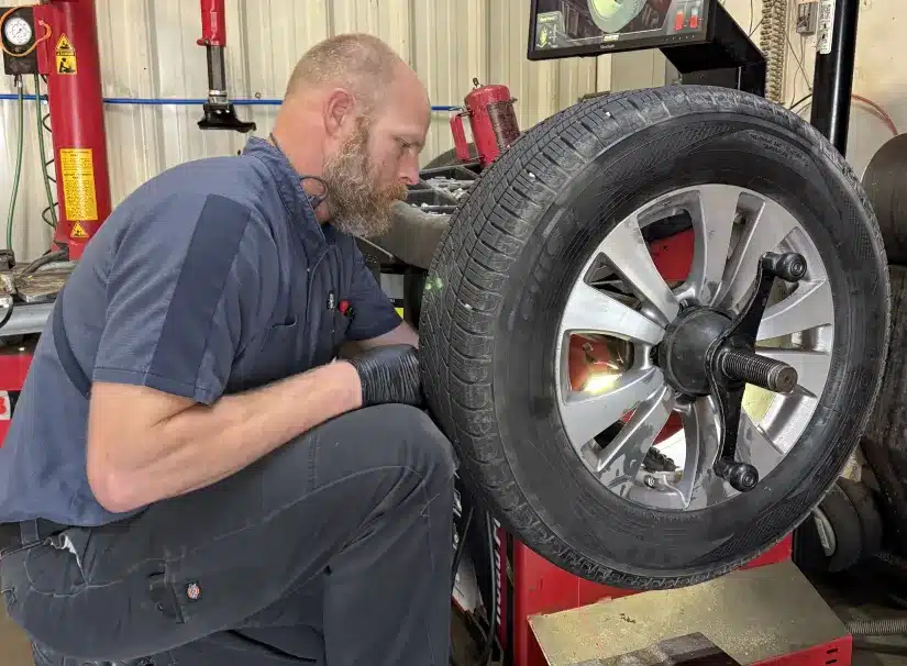 Tires Services at SPES Automotive. A mechanic with a beard and gloves is working on a car tire mounted on a machine inside an auto repair shop. He appears focused on inspecting or repairing the tire.