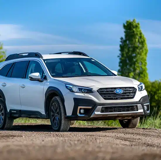 A white Subaru Outback SUV is parked on a dirt road with greenery and a clear blue sky in the background.
