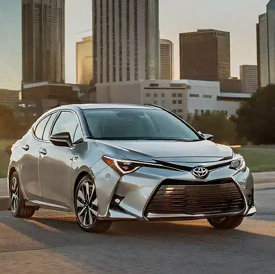 A silver Toyota sedan is parked on a city street at sunset, with tall buildings and a clear sky in the background.